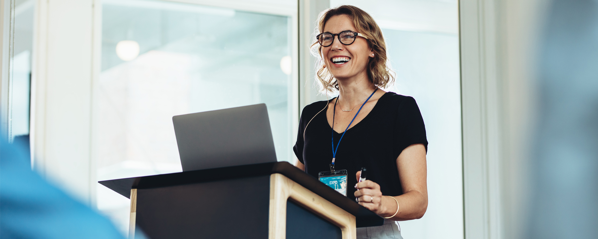 junge blonde Frau mit Brille steht lachend an Rednerpult, aufgeklappter Laptop für Präsentation steht am Pult