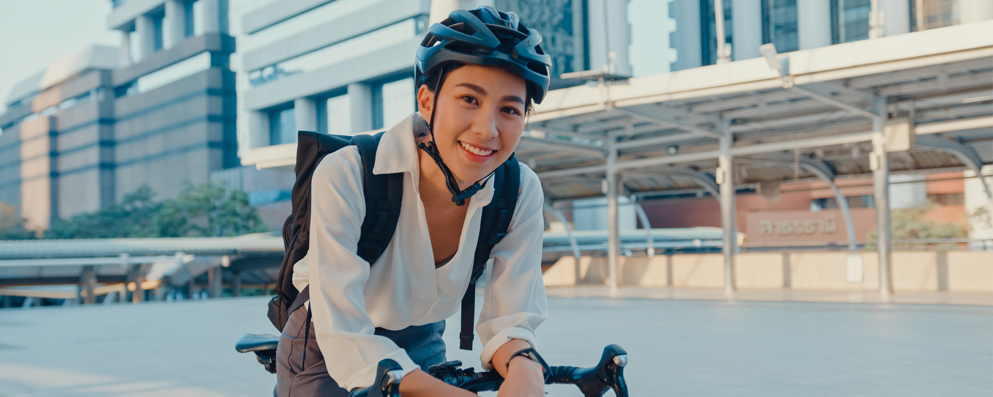junge Frau im Businessoutfit und Radhelm lehnt sich an Lenker und lächelt. Firmengebäude im Hintergrund